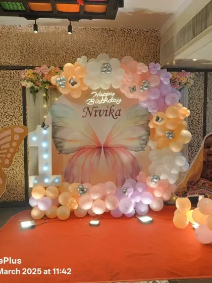A lovely butterfly-themed setup for Nivika's first birthday. The decor features a large butterfly wing backdrop, a circular balloon garland, and a smiling birthday girl enjoying her special seat.