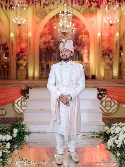 A full-length portrait of the groom standing proudly in his white sherwani in front of the wedding stage.