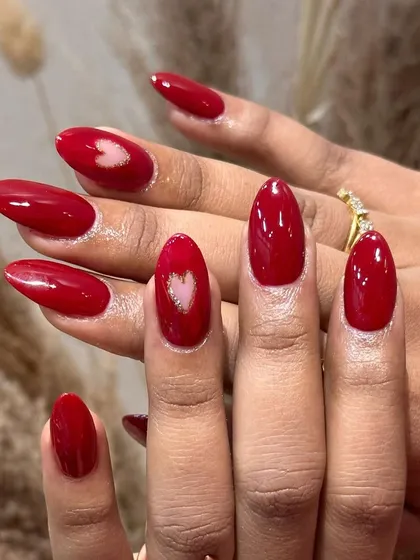 A full view of this romantic red manicure. The almond-shaped nails are painted in a vibrant red, with two accent nails featuring a beautiful, shimmering heart design.