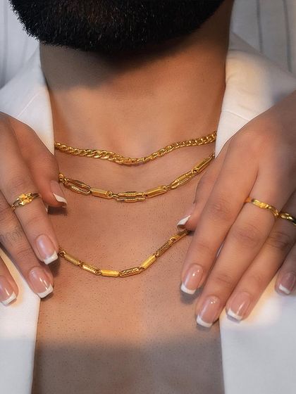A close-up showcasing the art of layering with our Noya collection, where delicate women's rings adorn hands that frame our modern men's gold chains.