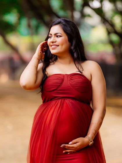 A beautiful, candid shot of a mom-to-be in a red gown, looking off to the side with a happy smile. The natural lighting and outdoor setting highlight her pregnancy glow.