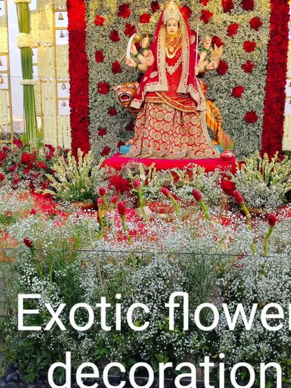 A grand outdoor setup for a Durga Puja or Navratri event. The goddess idol is placed before a massive backdrop of red roses, surrounded by a field of baby's breath.