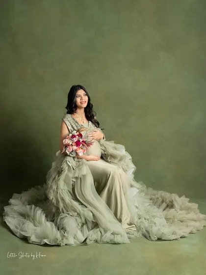 A seated portrait of a mother-to-be in a magnificent ruffled gown. She holds a bouquet of flowers, looking up with a serene expression against an olive-green backdrop.