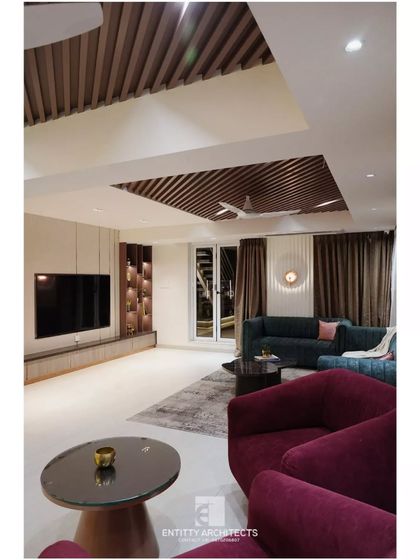 A full view of the living room, showcasing the interplay of textures and forms. The deep maroon armchairs add a pop of color against the neutral travertine wall, while the slatted wood ceiling adds warmth and architectural interest.