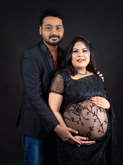 A classic studio portrait of a couple in traditional black attire. The sheer saree beautifully reveals the baby bump, blending tradition with a modern touch.