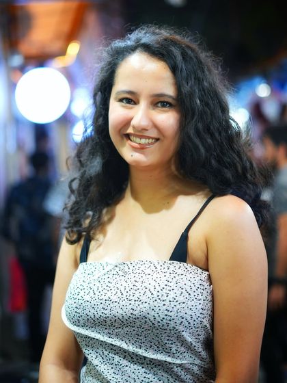 A warm, smiling portrait taken at night in Majnu Ka Tila. The soft bokeh from the lights in the background adds a magical touch to her friendly expression.