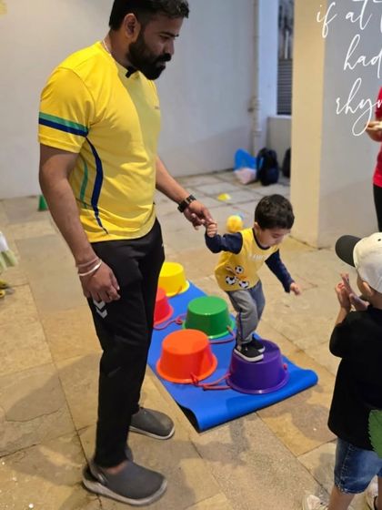 Here's a close-up of our bucket-walking exercise. A coach provides a steady hand while another child watches and claps, creating a supportive and encouraging atmosphere for learning new skills.