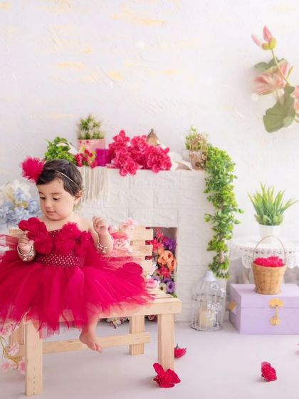 A little girl in a vibrant red tutu sits on a bench in a bright, floral-filled setting.