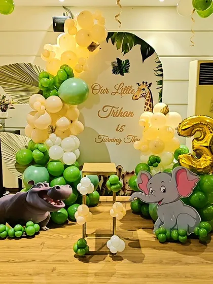 A cute jungle-themed decoration for a 3rd birthday. This setup includes a custom arched backdrop, elephant and hippo cutouts, and an organic balloon garland in green and yellow tones.