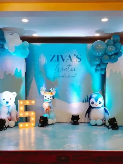 A cool "Winter Wonderland" themed 5th birthday party. The stage is decorated with blue and white balloons, cutouts of a polar bear, penguin, and reindeer, and a snowy backdrop.