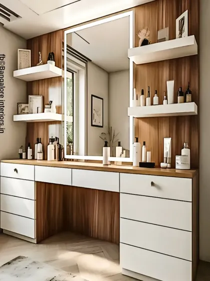 A modern dressing table with a large, backlit mirror providing perfect lighting. The unit offers extensive storage with multiple drawers in a clean white finish, contrasted with a wood-paneled wall and floating shelves for display.