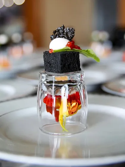 A deconstructed appetizer served in a unique way, with a savory black cube balanced on an inverted glass containing edible flowers. This is an example of our innovative and multi-layered plating style.