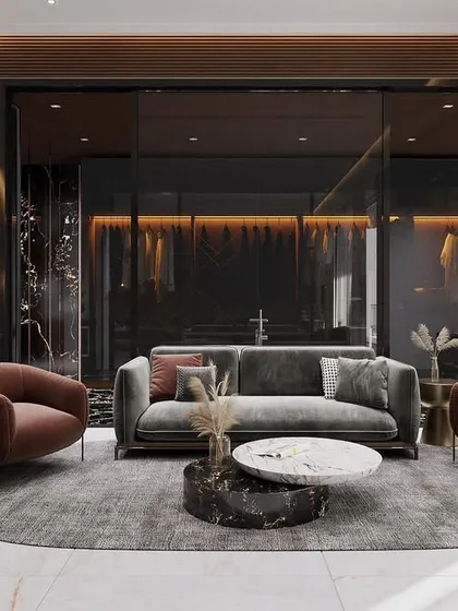 This seating area, part of a larger master suite, is designed for relaxation. It features plush, contemporary furniture in muted tones, a layered rug over polished marble flooring, and a view into the walk-in closet through smoked glass panels.