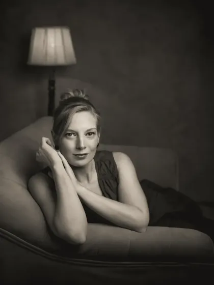 Sonia's portrait in monochrome takes on a quiet, contemplative quality. Leaning on the couch with a vintage lamp in the background, the scene feels intimate and cinematic. The black and white tones enhance the moody, soulful atmosphere of the shot.