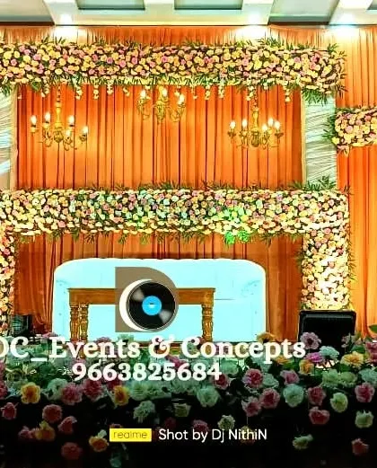 A grand, full-width stage decoration for a large auditorium wedding. The design features a warm orange and peach color palette with extensive floral work, transforming a simple hall into a spectacular wedding venue.