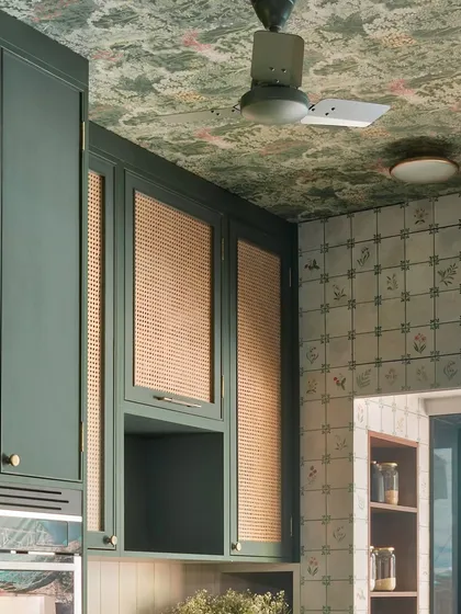 The ceiling wallpaper and patterned wall tiles create a rich tapestry of design in this kitchen. The wicker-fronted cabinets add another layer of texture, making the space feel deeply personal and curated.