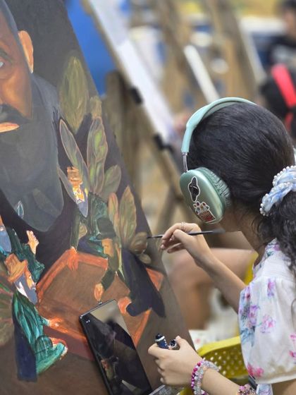 A young student, focused and in her own world with headphones on, carefully paints a portrait during class.
