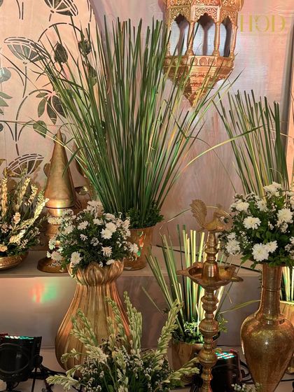 A close-up of a decor corner featuring traditional brass vases, a multi-tiered lamp, and lush greenery, set against a backdrop of lotus motifs.