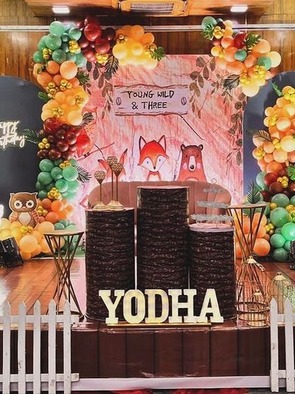 A whimsical woodland animal theme for a third birthday. This unique setup uses balloon trees, a picket fence, and cute fox and owl characters to create an enchanting forest scene on stage.