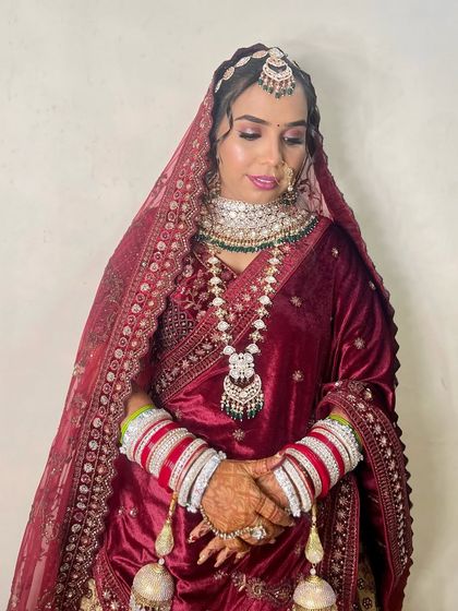 A bride in a deep red velvet outfit. Her hair is styled in a classic updo, allowing the heavy dupatta and jewelry to be the main focus.