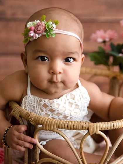 A baby girl in a lace romper sits in a wicker basket, looking at the camera.