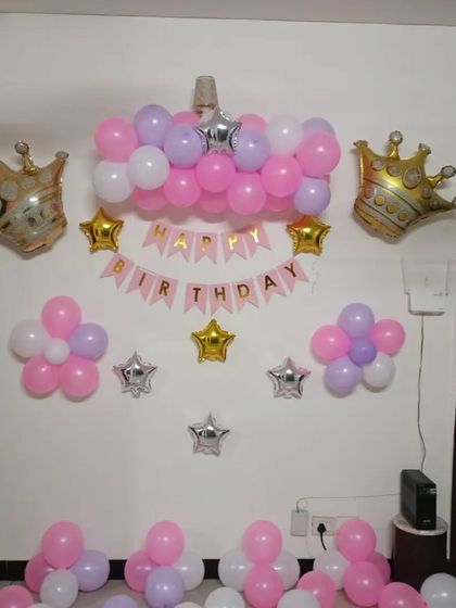 A simple princess-themed wall decoration for a home party. The setup includes a 'Happy Birthday' banner, foil crowns, and balloon clusters in pink, purple, and white.