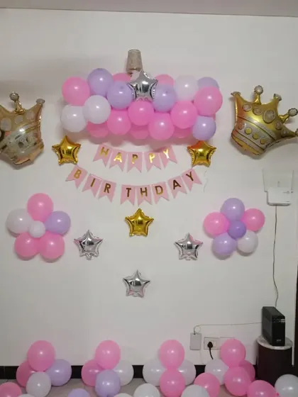 A simple princess-themed wall decoration for a home party. The setup includes a 'Happy Birthday' banner, foil crowns, and balloon clusters in pink, purple, and white.