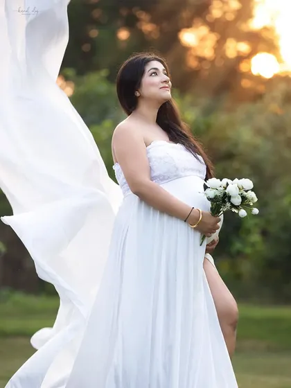 An outdoor maternity portrait taken during the golden hour, with soft sunlight creating a warm, ethereal glow. The flowing white gown and natural setting provide a romantic and serene feel.