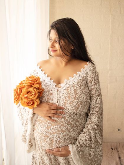 A beautiful solo portrait of a mother-to-be in a delicate lace robe, holding a bouquet and looking out the window.