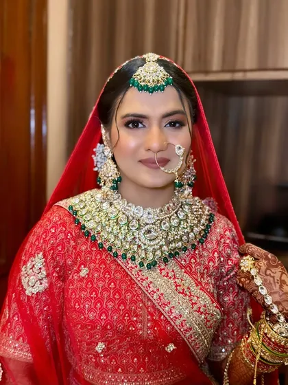 Royal hues for a graceful bride. This look features a stunning red lehenga with green jewellery, and the makeup is balanced to complement both colors perfectly.