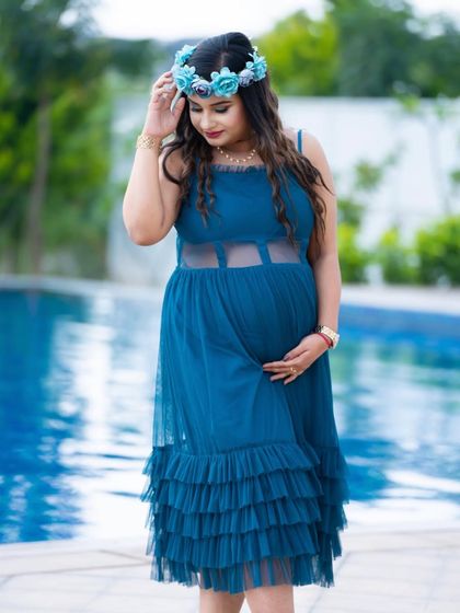 A serene moment by the water. The mom-to-be, in a blue dress and floral crown, stands by a pool, creating a calm and beautiful portrait.