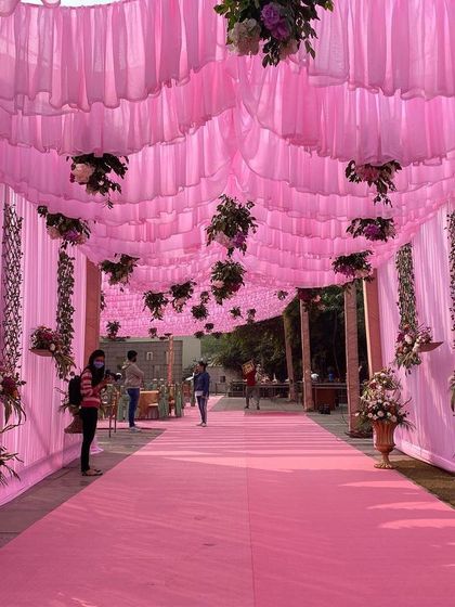 The entrance walkway for a daytime event, decorated with a ceiling of pink drapes and hanging floral arrangements, creating an immersive and colorful welcome for guests.