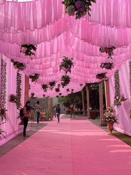 The entrance walkway for a daytime event, decorated with a ceiling of pink drapes and hanging floral arrangements, creating an immersive and colorful welcome for guests.