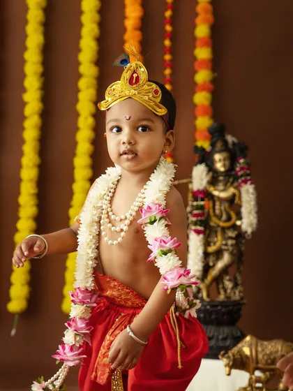 A solo portrait of a toddler dressed as Krishna, standing confidently in front of a deity statue.