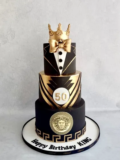 A cake fit for a king's 50th birthday. This three-tiered design in black and gold features a tuxedo on the top tier, a Versace-inspired Medusa plaque, and a golden crown.