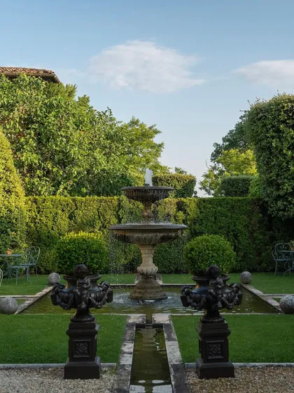 We create classic, formal gardens that evoke timeless elegance. This space is defined by its perfect symmetry, featuring a central tiered water fountain, meticulously manicured topiary, and lush green lawns bordered by high privacy hedges.