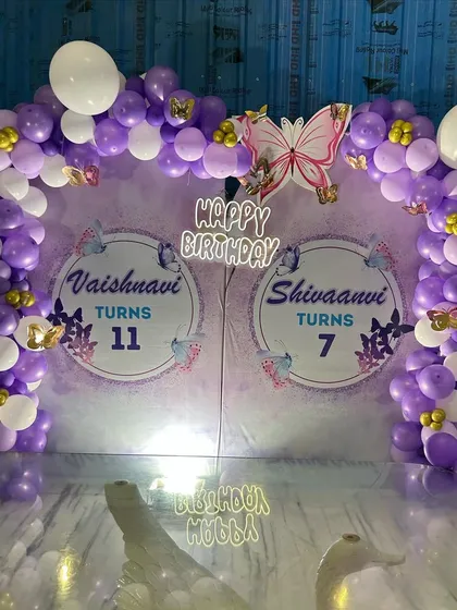 A shared birthday celebration with a purple and white butterfly theme. The backdrop is customized for two birthdays and is framed by a matching balloon arch.