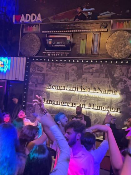 A shot from below, looking up at the iconic DJ booth at Bombay Adda, with the crowd's hands in the air.