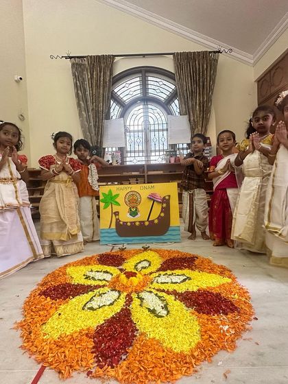 Children stand in a respectful 'namaste' pose around the completed pookalam, a beautiful culmination of their collective effort.