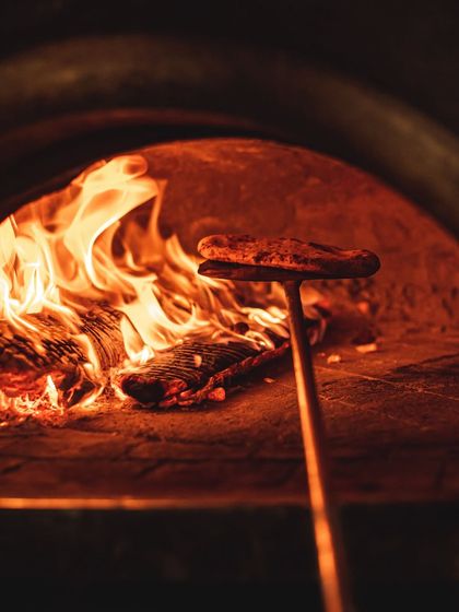 A pizza peel holds a baking pizza close to the burning wood, a technique called 'doming' that helps cook the toppings and melt the cheese perfectly.