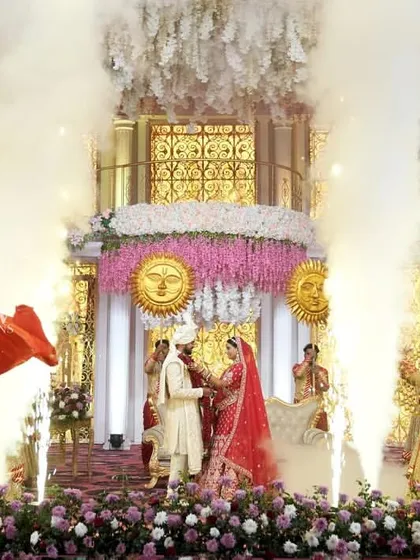 The Jaimala ceremony is elevated with dramatic cold-fire pyrotechnics and smoke effects. The couple stands on a beautifully decorated stage, surrounded by dancers and flags, creating a powerful visual.