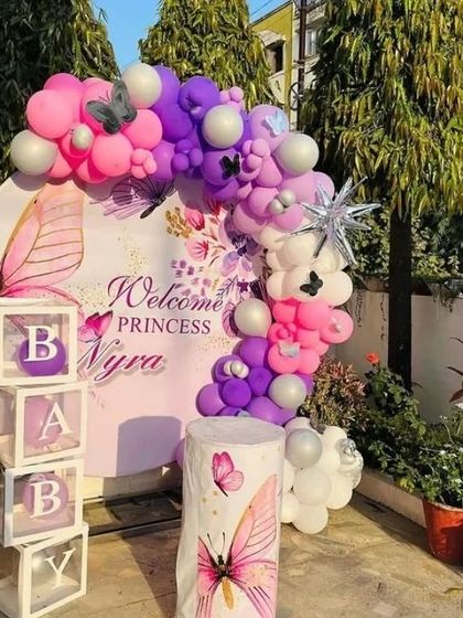 An outdoor welcome for a princess, featuring a personalized circular backdrop against a backdrop of lush greenery. The setup includes "BABY" blocks, a themed pedestal, and a beautiful balloon garland in pink, purple, and white.