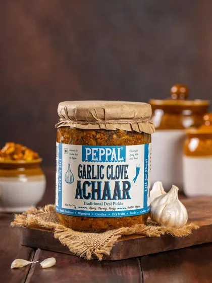 A product shot of Peppal's Garlic Clove Achaar, styled with fresh garlic and rustic pottery. The composition reinforces the traditional, flavorful nature of the pickle.
