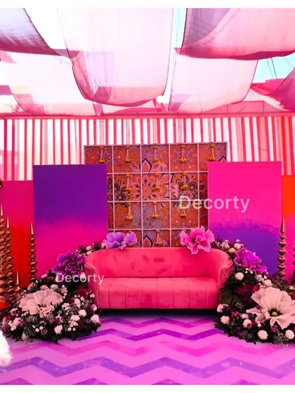 A vibrant and colorful Mehendi ceremony stage, combining bright pink and purple drapes with lush floral arrangements. The setup is designed to create a festive and traditional 'mehendi waala feel' for the celebration.