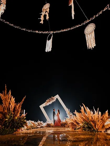 A breathtaking wide shot of an engagement setup in Goa. The couple stands framed within a large white structure, surrounded by pampas grass and the dark night sky, creating a dramatic and romantic scene.