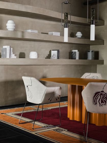 A dining or meeting space set against a wall of built-in shelves. The embroidered chairs and bold orange table create a strong focal point, while the neutral, lime-washed shelves provide a calm backdrop for decorative objects.