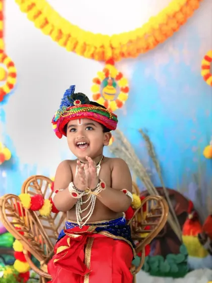 Capturing the joyful spirit of Janmashtami with a toddler dressed as Krishna, clapping his hands with a wide smile. The festive backdrop is decorated with marigold garlands.