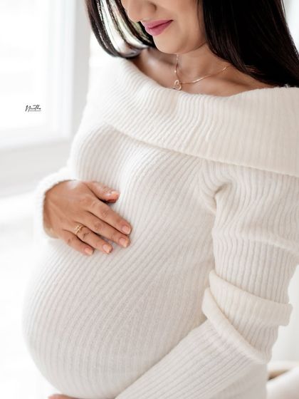 A close-up detail shot of a pregnant mother's hands on her belly, wearing a beautiful white ribbed dress.