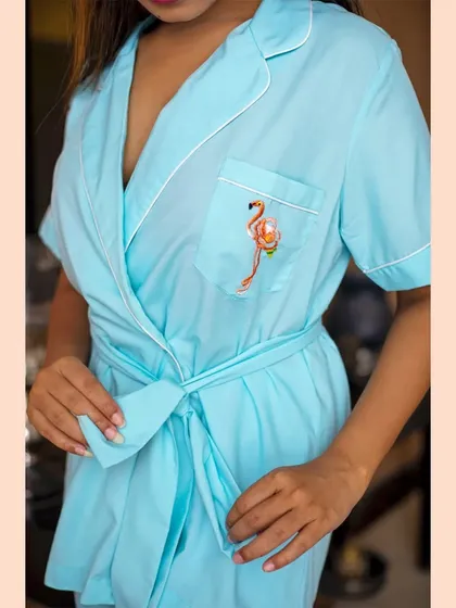 A detailed shot focusing on a light blue robe. The image highlights the custom flamingo embroidery on the pocket and the crisp white piping, showcasing the brand's attention to unique, handcrafted details.