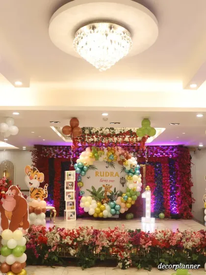 A wide-angle view of the entire stage, showing the grand scale of the jungle decoration. The combination of the chandelier, flower-draped ceiling, and the themed setup creates a truly luxurious and memorable first birthday celebration.